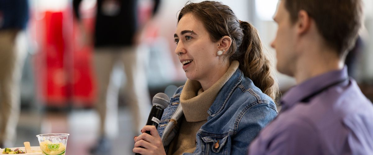 Member of the faculty and staff speaking at a luncheon holding a microphone with people seated around them