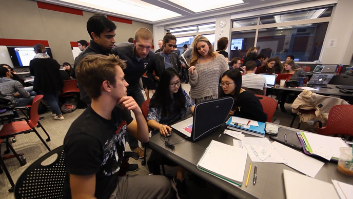 A group of 7 BU students work together as one student types at a laptop and the rest look over her shoulder at the screen.