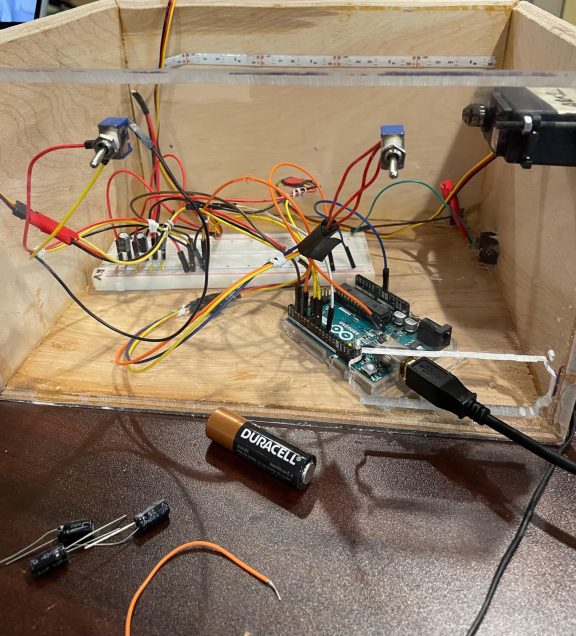 a wooden box that is open on top with a wall of plexi as the front of the box. Inside the box are a variety of electronic components, some of which are coming out of holes in the plexi as well