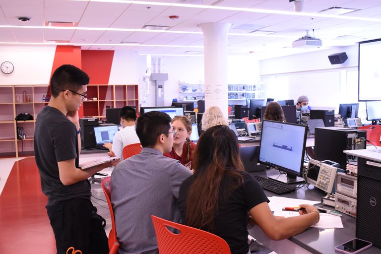 a group of 4 students working at one of the computers in the dry lab at BTEC. 3 are sitting, 1 is standing and holding a closed laptop