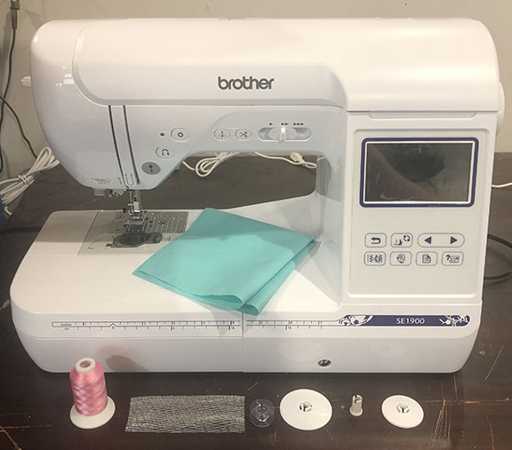 the brother SE1900 seiwng and embroidery machine, turned off with a spool, empty bobbin, spool net, and spool caps on the table in front of the machine. There is a square of layered blue fabric in the throat of the machine