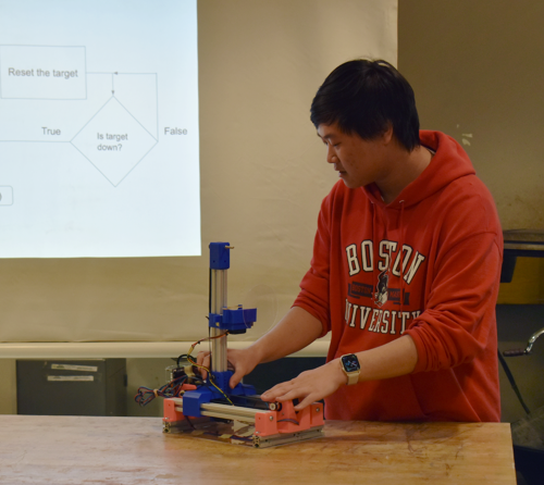 male student in red BU hoodie demonstrating how a project made of 3D printed parts and aluminum bars works in front of a projector screen with technical details about the project