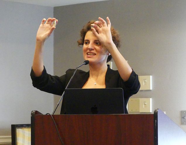 Professor Yazicigil behind a podium, holding up a tiny item between the thumb and forefinger of each hand.