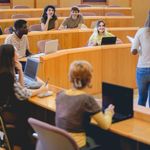 A lecture hall filled with people sitting in chairs, engaged in a presentation by a speaker at the front.