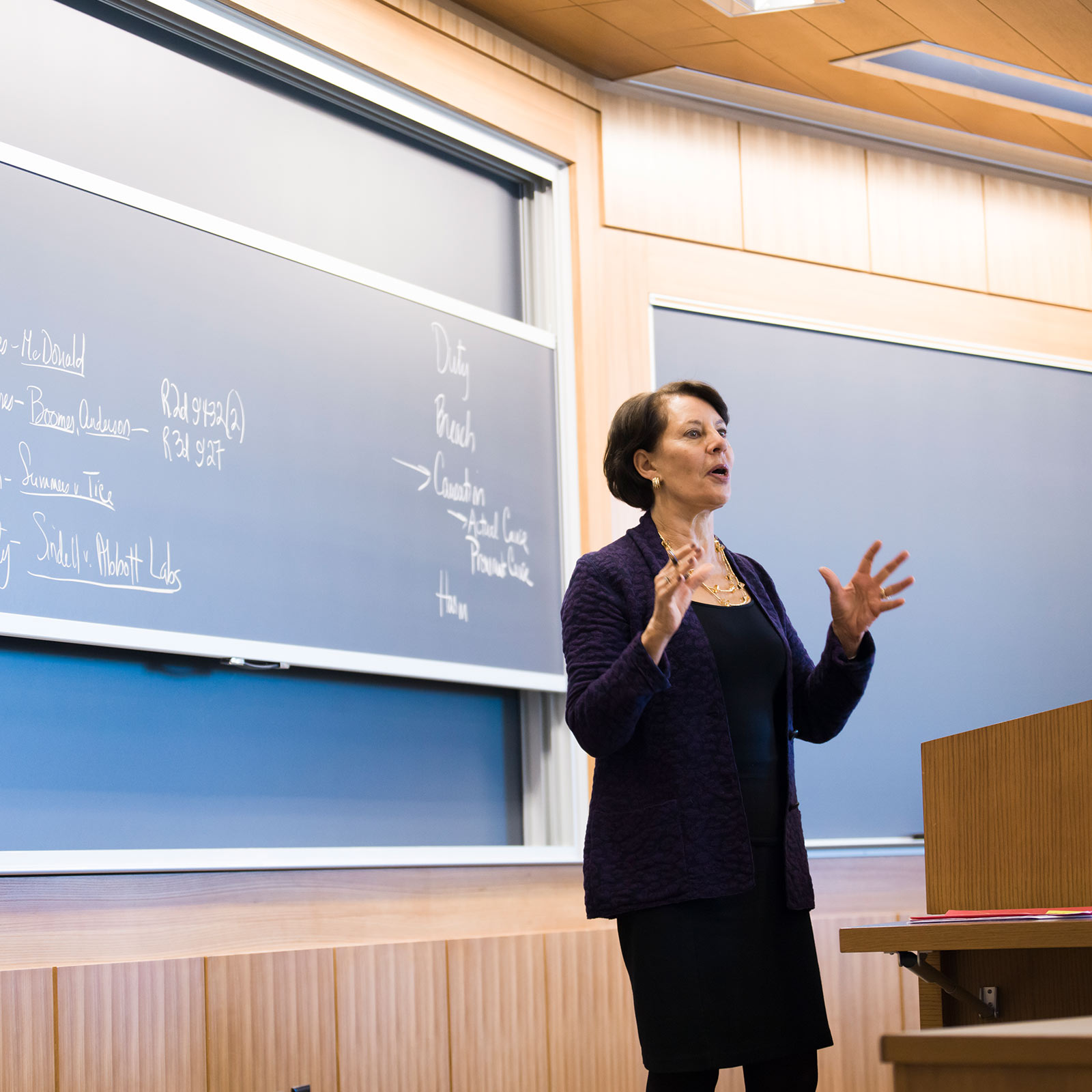 A class is in session at the BU School of law. The professor is a mature woman wearing professional clothes that are all black, which matches her parted dark hair. She appears mid-sentence with hands gesturing an explanation of a concept.