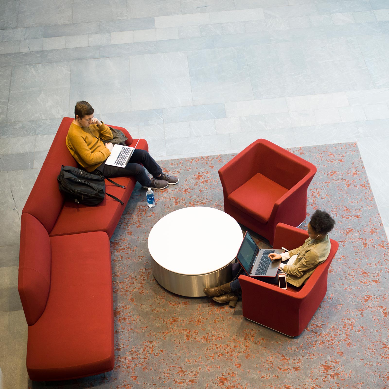Two students are studying in a well-lit lobby of BU's LAW school building. They appear to be wearing warm clothes, suggesting a fall or winter. The chairs are comfy couches with a BU Red color, while a metallic circular table appears in the middle.