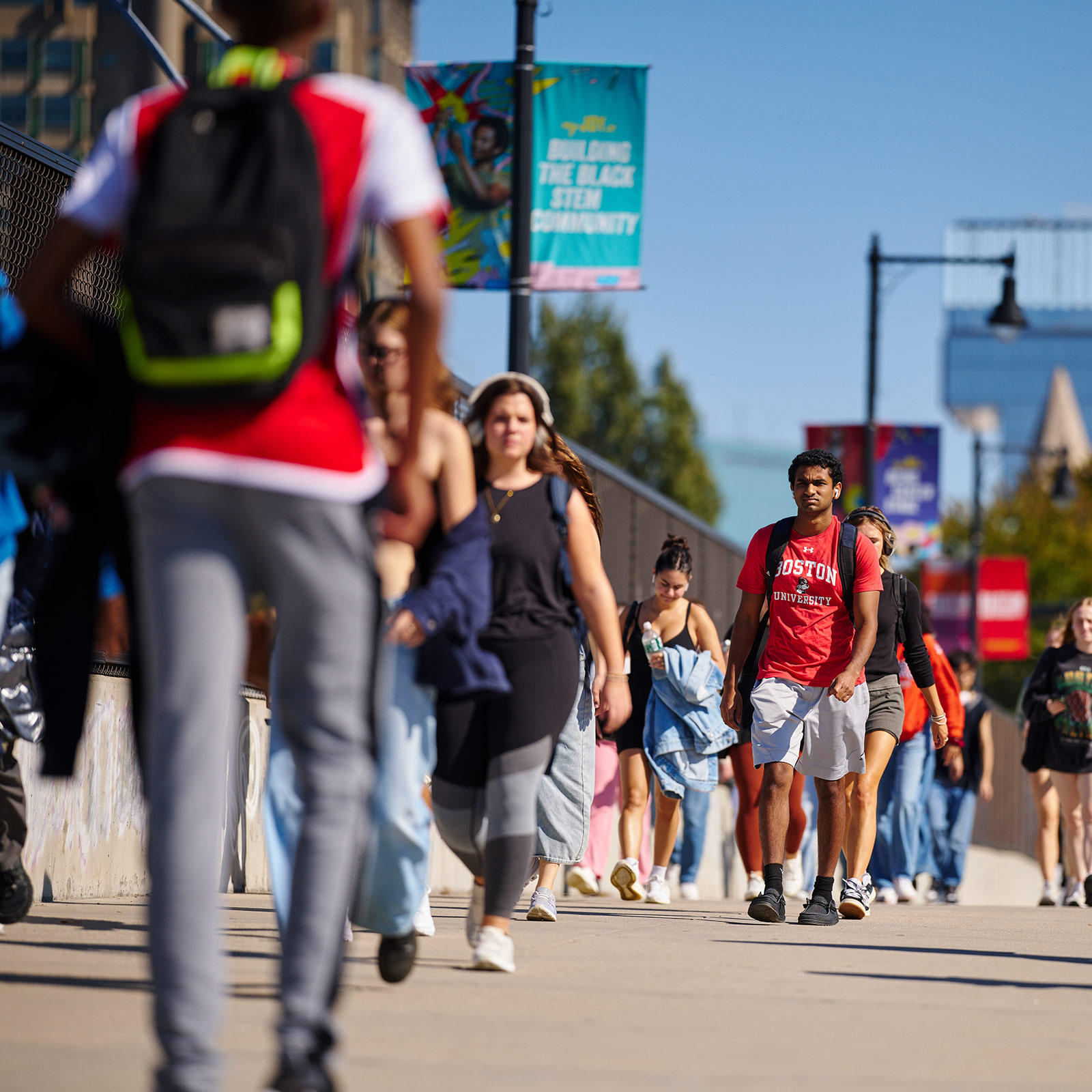 A mass of students on a spring day are commuting along Commonwealth Avenue between the BU Bridge and the CFA building to class. Some are wearing BU-branded T-shirts in the University red color. almost everyone has a focused look.