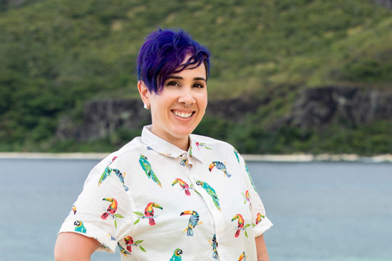 幸存者 contestant Lyrsa Torres and BU alum poses on a white sandy beach in a button down shirt with colorful Toucans