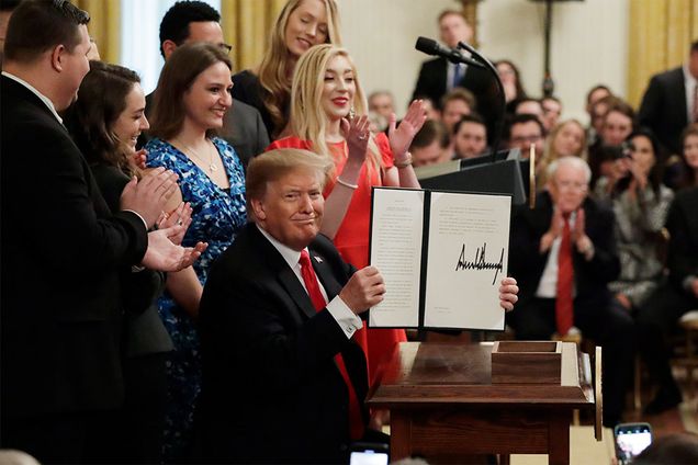 President Donald Trump holds up an executive order on campus free speech, surrounded by college students.