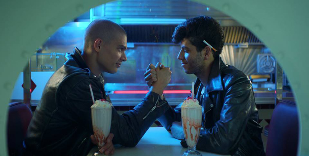 A gay couple holds hands while sitting at a table with milkshakes.