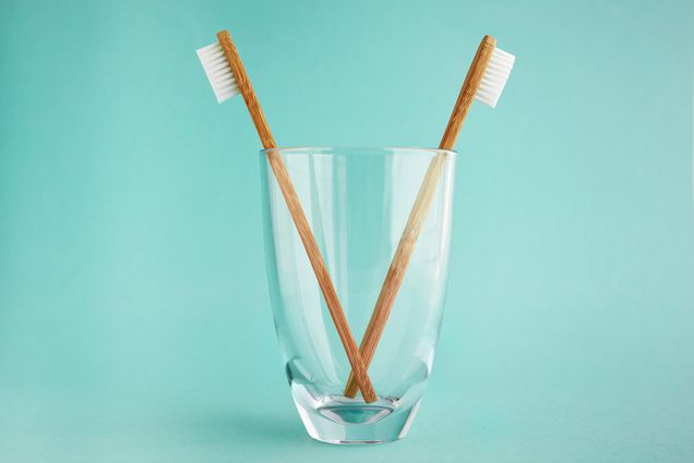 Two bamboo toothbrushes in a water glass over a seafoam green-blue background