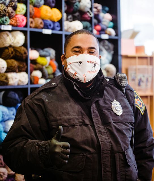 A police officers gives a thumbs up while wearing a cloth face mask with Rhett and the words "Boston University" on it. The officer stands in the Stitch House in Dorchester.