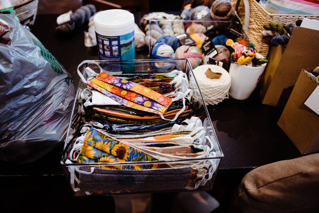 Image of a pile of hand-sewn, brightly-colored cloth face masks inside a plastic tub, on top of a table with other crafting supplies.