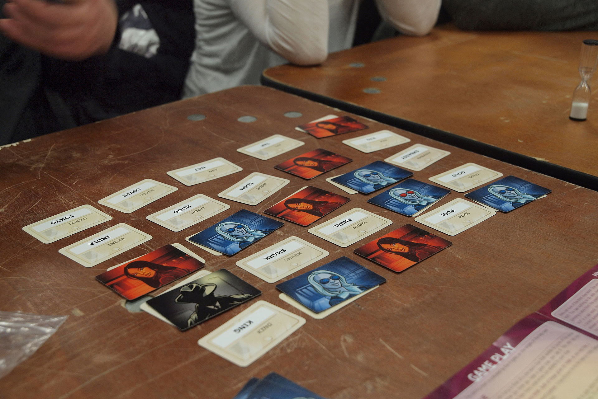 Photo of a game of Codenames being played at the annual board game festival of Finland in Helsinki, Finland. Some cards in the five by five grid show words, while others have blue, red and black illustrations of people.