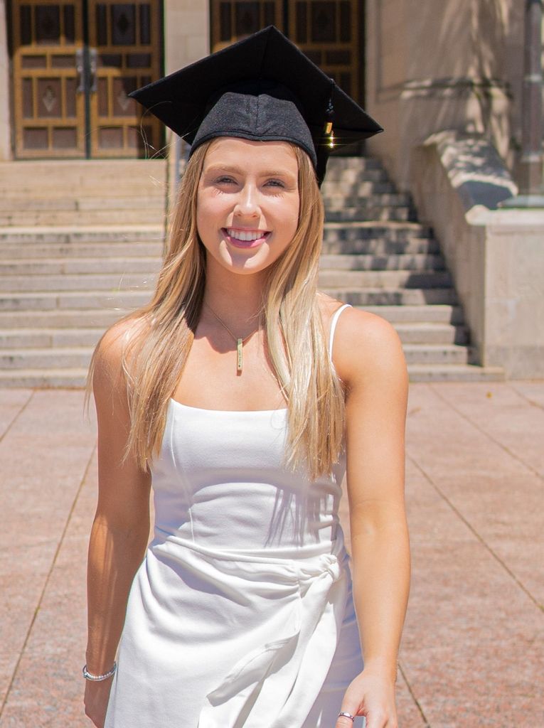 Photo of McKenzie Beaton (SAR’20) in a white dress and black graduation cap in front of Marsh Chapel.