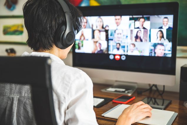Photo of a student with headphones looking at a desktop screen with a video chat call is seen. The student's hand is on the mouse.