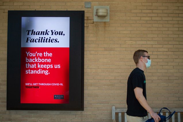 A person in a mask walks down Commonwealth Ave on June 16, 2020. A sign on the wall behind them reads: "Thank You, Facilities. You’re the backbone that keeps us standing. We’ll get though COVID-19 together." Boston University logo is seen in black on the bottom right corner.