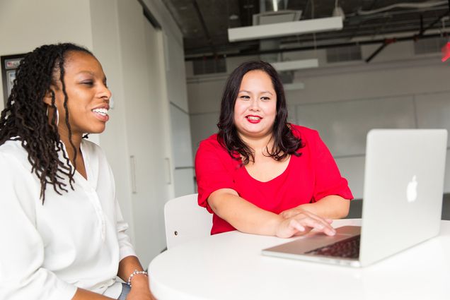 Two women look at a laptop screen, one smiles and wears a white blouse, the other, in a red blouse, types on the keyboard.