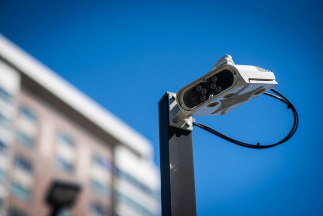 New cameras are seen at the Agganis Arena parking garage on August 3, 2020. The new parking program will read license plates instead of stickers or hang tags. A camera with orange lights hands next to a parking lot arm. Blue sky is seen in the background.