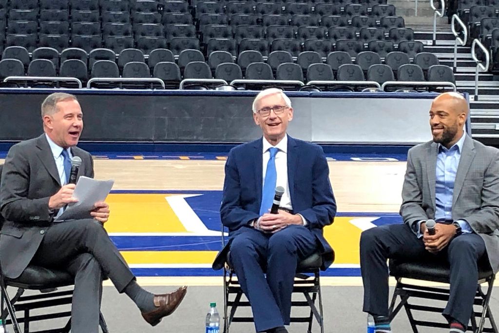 Wisconsin Governor Tony Evers (center) and Lieutenant Governor Mandela Barnes (right) take questions from Democratic National Convention CEO Joe Solmonese on January 7, 2020, on the floor of the Fiserv Forum in Milwaukee. The men wear suits, sit in folding chairs and stadium seating is seen behind them.