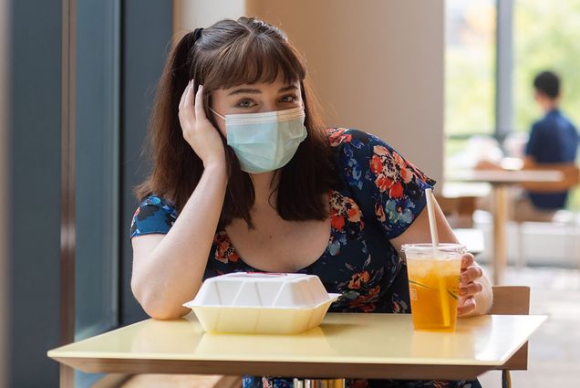 Photo of Brigid Kane (COM’22) sitting at a small dining hall table by herself with a face mask on. A paper to-go container and drink sit in front of her; she rests her hand on her face. Overlay has a video play button.