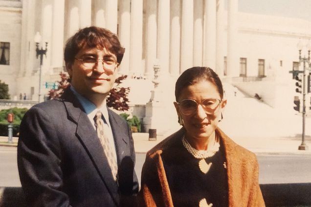 Photo of a printed photo of Jay Wexler (left) with Supreme Court Justice Ruth Bader Ginsburg in 1999, when he was one of her four clerks. Behind them is the facade of the Supreme Court. RBG wears a mustard colored shawl and Wexler wears a suit and tie.