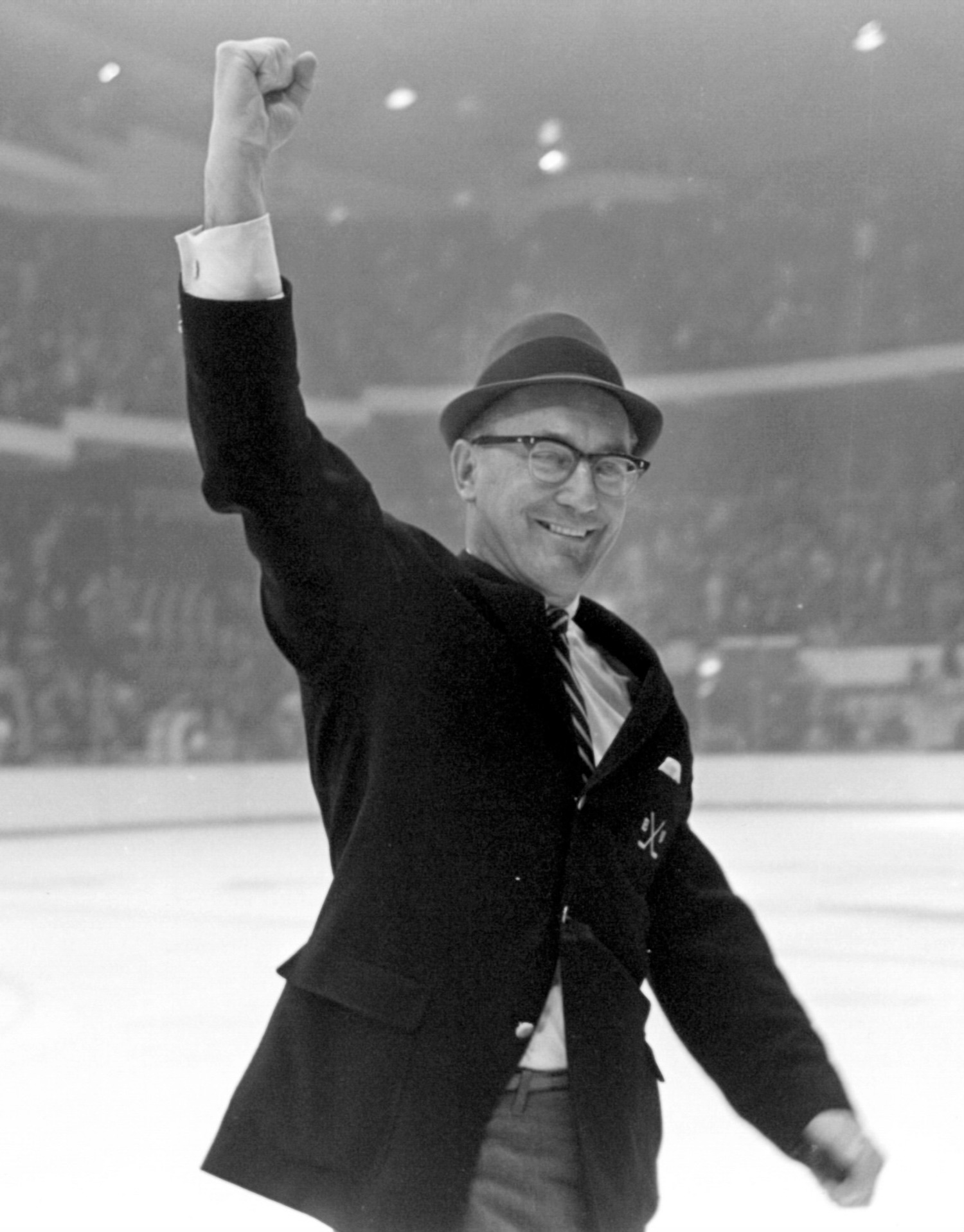 Black and white photo of Jack Kelley with his hand in air on the ice of a hockey rink. He wears a hat and glasses and smiles.