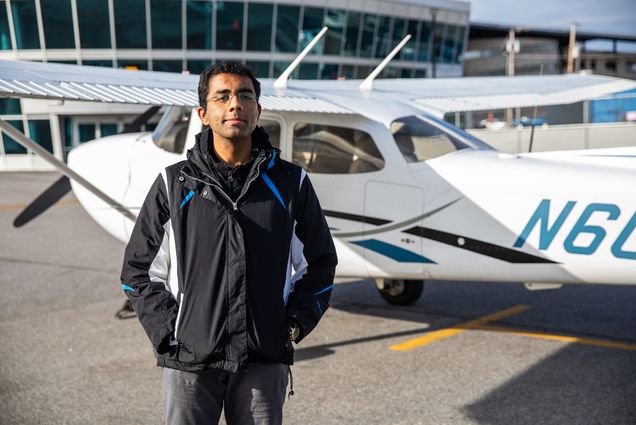 Photo of pilot Dharmesh Tarapore (CAS’18) standing in black jacket in front of a small plane and smiling.