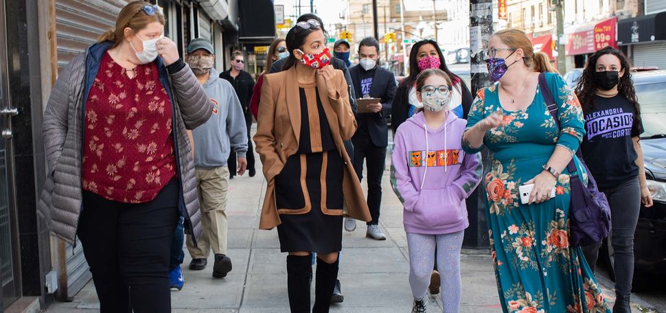 Photo of Alexandria Ocasio-Cortez (CAS’11) (D-N.Y.) campaigning in Queens, N.Y., on October 18, 2020. She wears a red mask and talks to a woman and a young girl to her left.