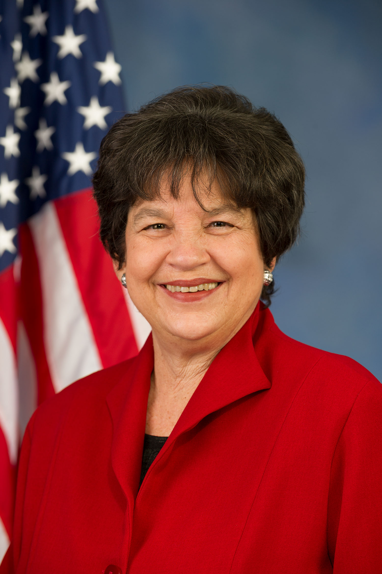 Portrait of Representative Lois Frankel (CAS’70), who is seeking a fifth term in Congress from Florida. She wears a red blazer, smiles wide, and an American flag is seen in the background on the left.