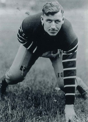 Black and white photo of Harold “Spike” Carlson (Questrom’26), in an old-fashioned football uniform touching the ground while squatting.