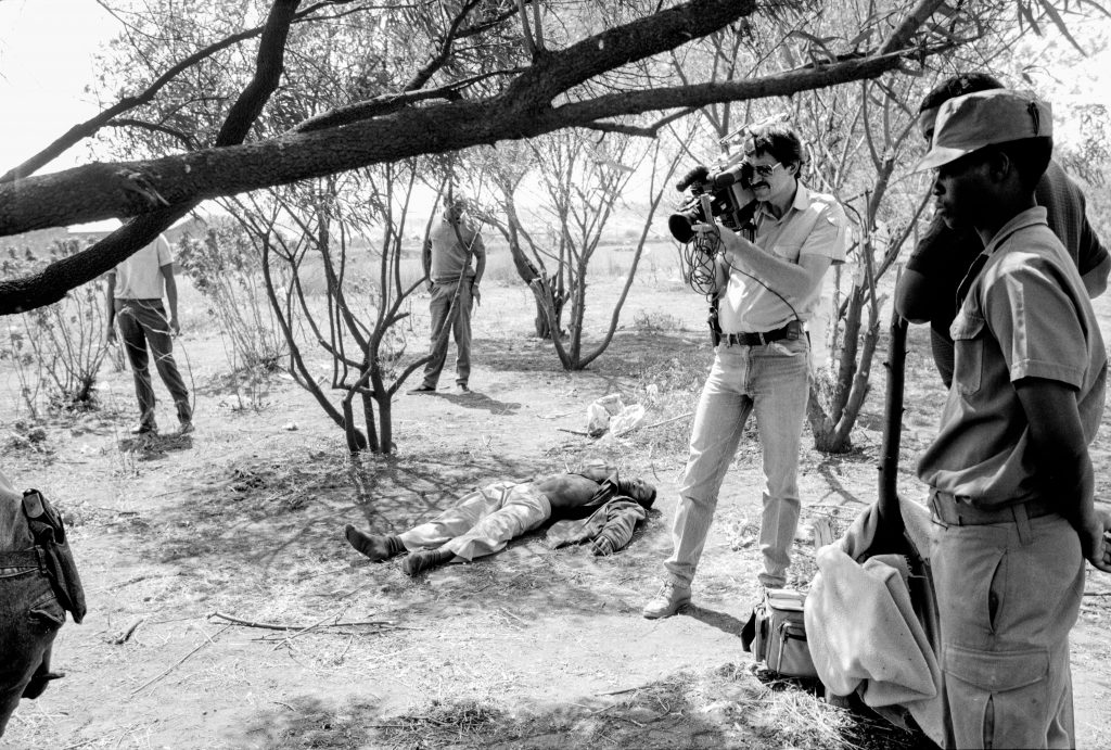 Black and white photo of a policeman with a video camera ignores the dead man near him following clashes in September of 1990 near Soweto’s Jabulani migrant workers hostel in clashes between African National Congress and Inkatha Freedom Party supporters.