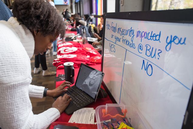 Photo of Michelle Njoroge (CAS,SAR'21), Director of the Department of Social Advocacy for the Student Government during a tabling event in which the group got the word out about Terrier Meal Sharing. Mjoroge sets-up the sign-up system on a laptop at a booth, a white board on the table reads “Has food insecurity impacted you or your friends at BU? Yes or No” with columns for responses.