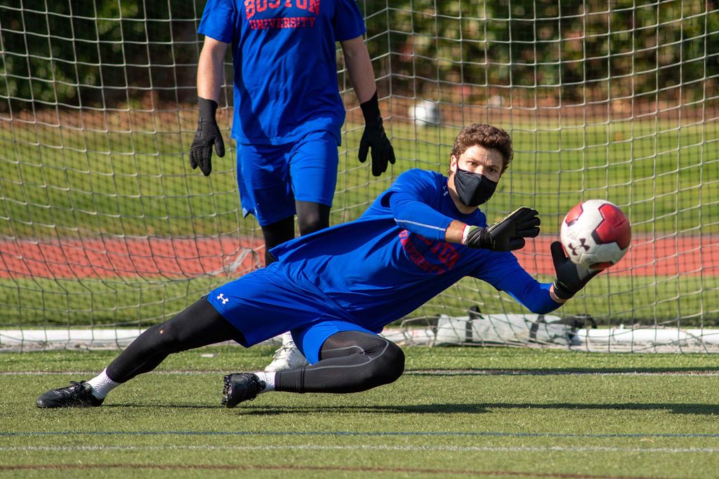 Photo of Michael Stone (CAS’21) during soccer practice. He wears a blue shirt and shorts and a face mask as he plays goalie and blocks a soccer ball.