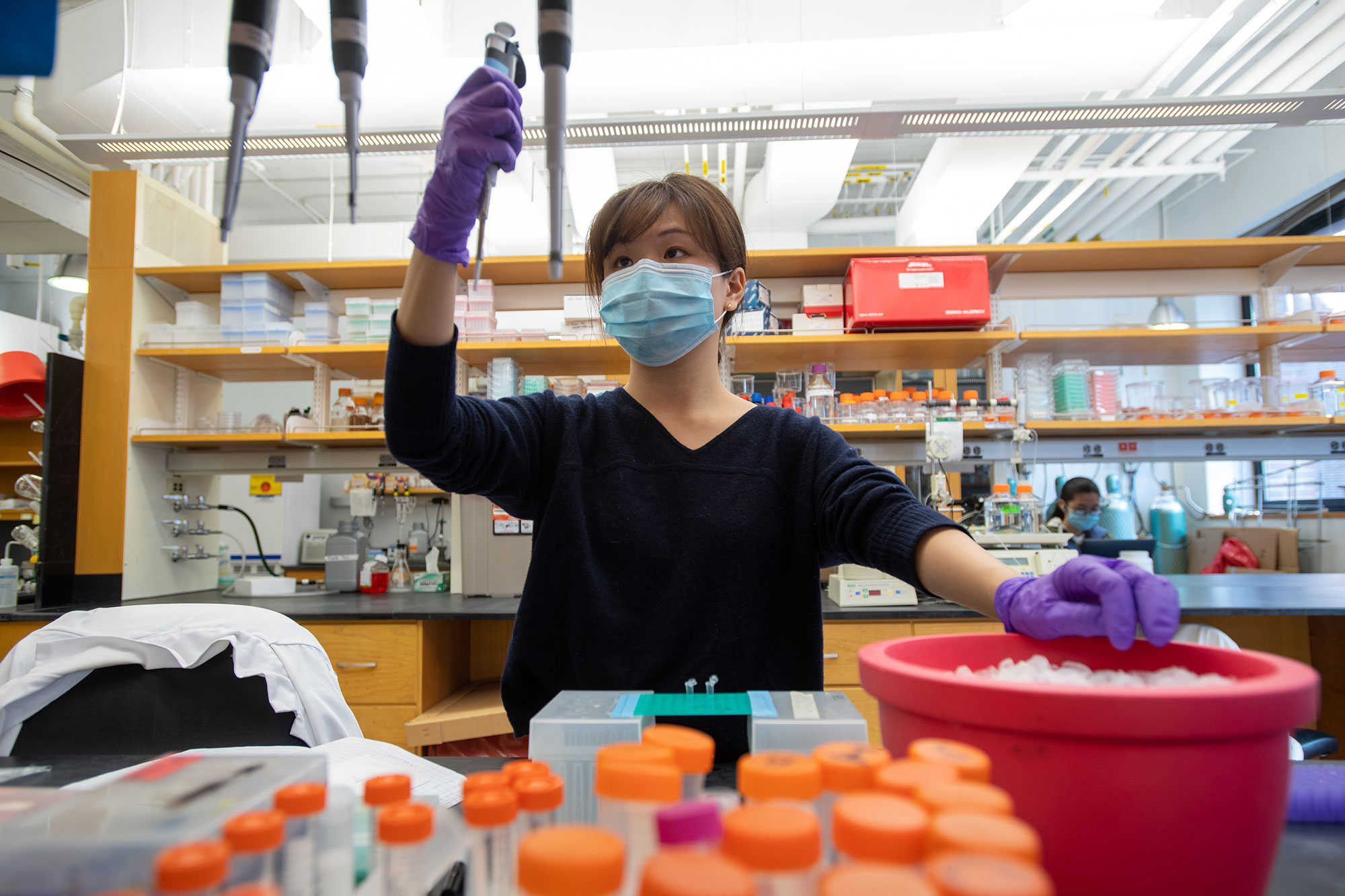 Photo of of Sunnie Kong (CAS'21, GRS'21) in a lab with a blue face mask on a she holds up a vile. She wears purple gloves. A dozen orange-capped vials are seen in the foreground.