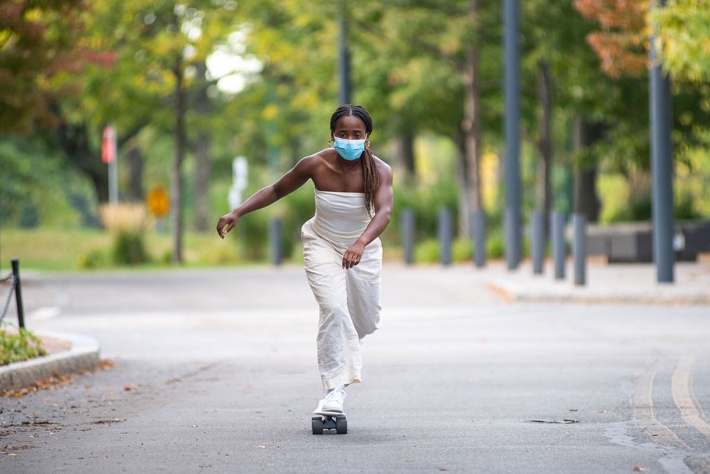 Photo of Archelle Thelemaque (COM’21) dressed in a white one piece and wearing a blue face mask skate boarding.