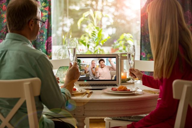 Photo of family members video chatting during a socially-distant holiday meal. The two people in the foreground hold champagne flutes and have plates of food in the front of them. On the laptop screen, two people, presumably relatives, also hold up wine glasses, as if making a toast.