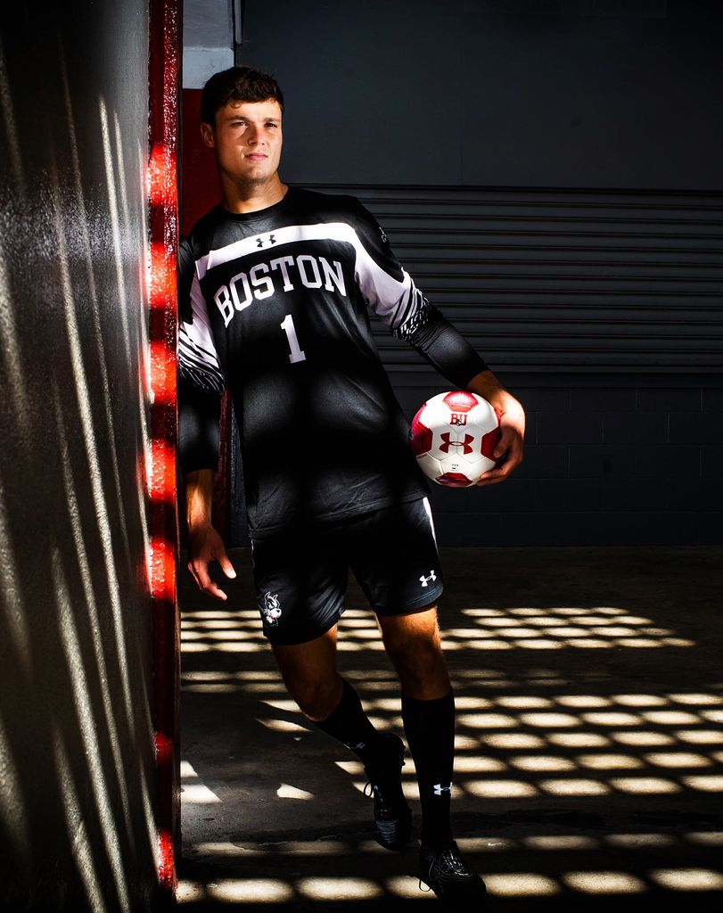 Photo of Michael Stone (CAS’21)in a dimly lit locker room with light coming through in a square grid. He wears a black BU jersey with the number one and holds a red and white soccer ball to his side.