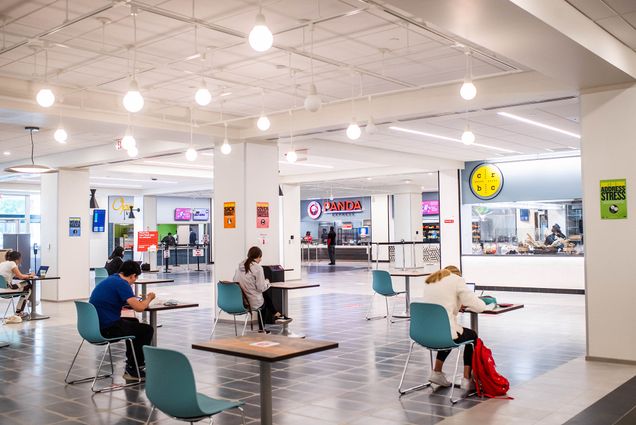 Photo of the recently renovated GSU Food Hall. Single person tables with teal seats are spread out in the foreground, a few students sit and eat. In the background, the white lights and walls and a few food stalls are seen.