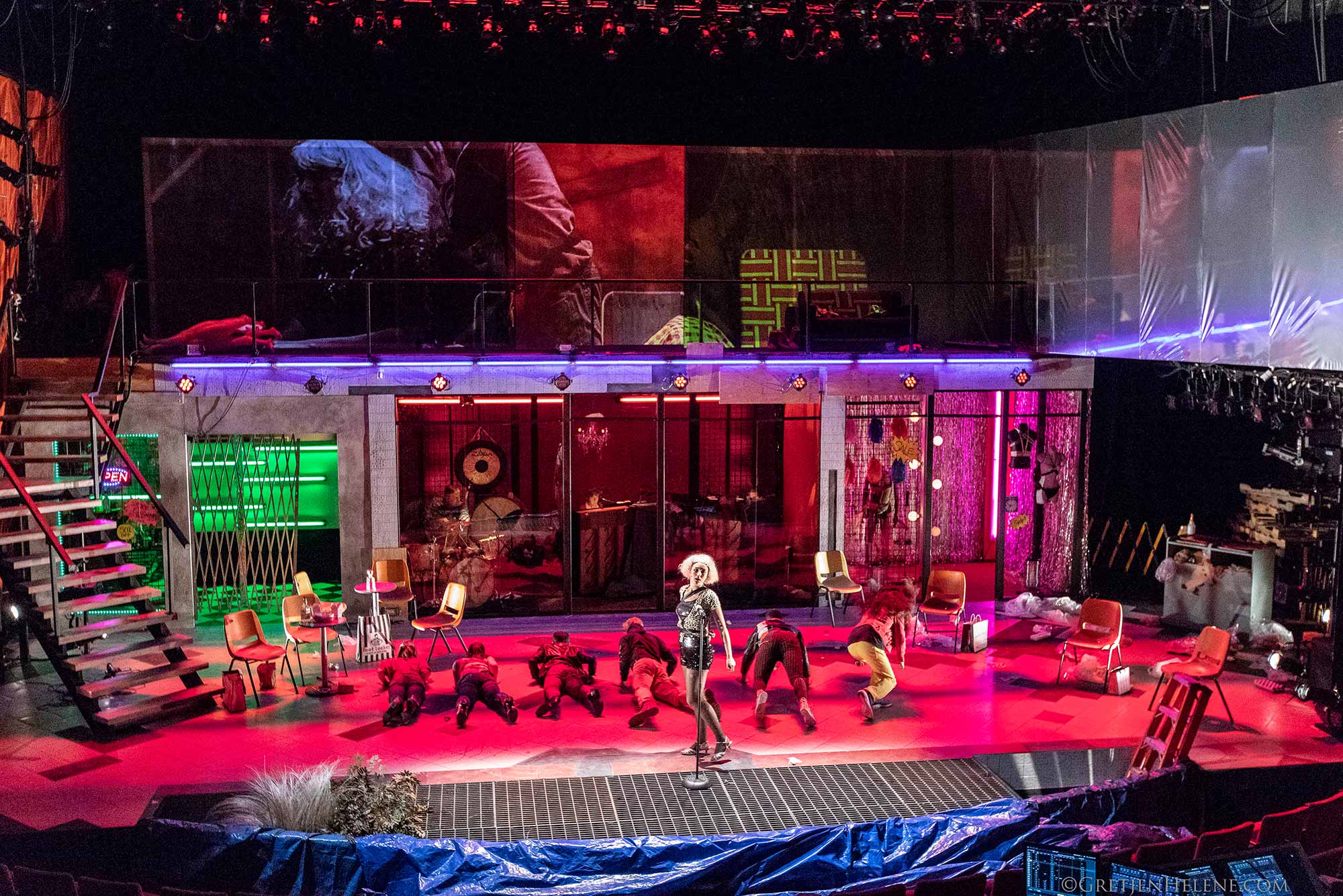 Photo of a set in 2017 production of Cabaret that Pajoufar worked on. It is brightly lit with pink and purple lighting. One person stands center stage while a group of people behind them lay flat on their bellies facing away from the audience. The set looks like the inside of a shop.