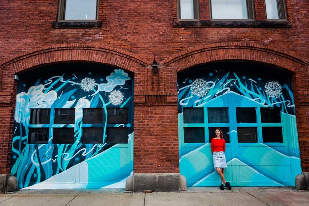Photo of muralist Lena McCarthy standing in a red long sleeve shirt and jeans in front of a pair of doors on a Victorian firehouse that she painted with a large mural of various shades of blue.
