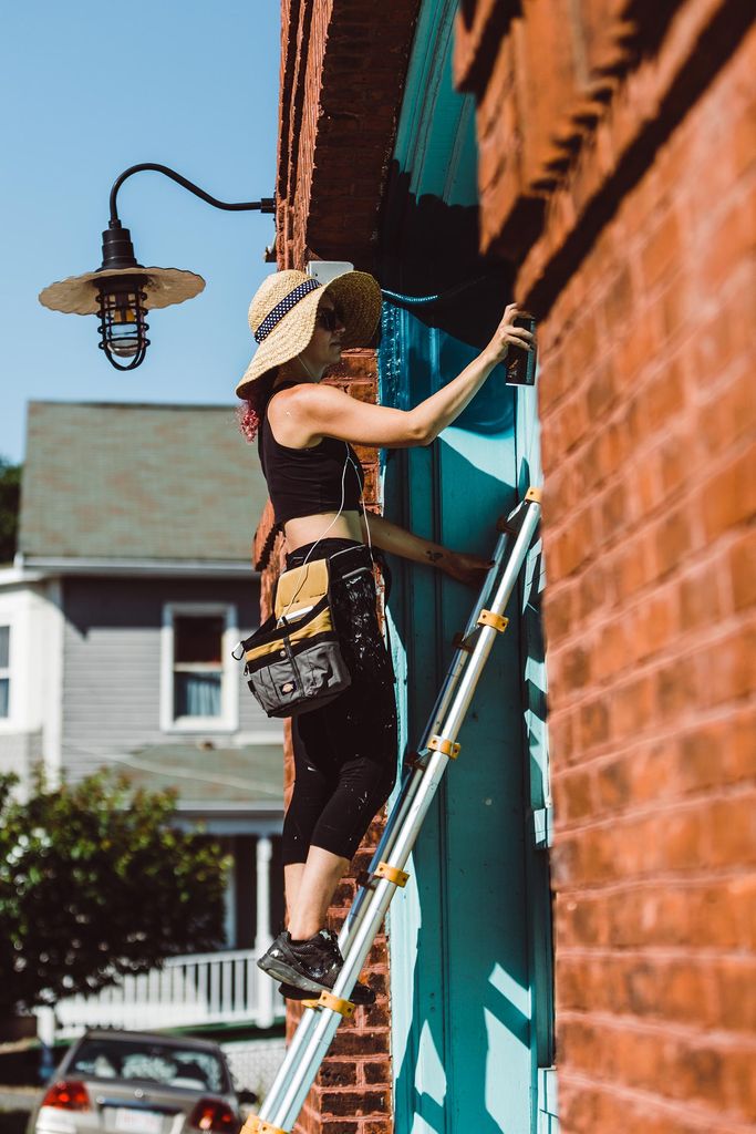Photo of muralist Lena McCarthy from the side. She is standing on a ladder in sunglasses, a large sun hat, and tool belt with a spray paint can in her hands as she works on the mural on the firehouse doors.
