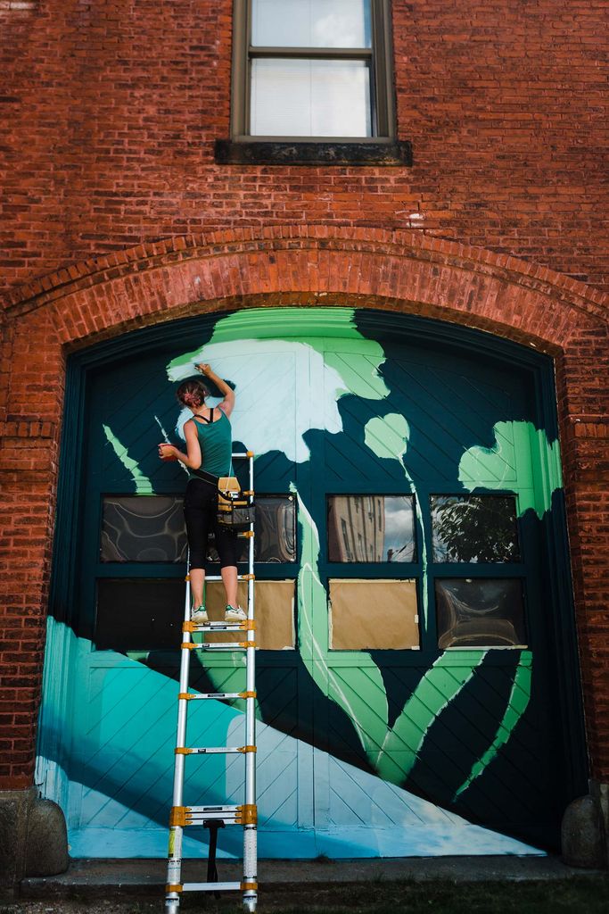 Photo of Lena McCarthy in black paints and a green shirt, standing on a ladder as she works to paint one side of the doors. The mural depicts forms that resemble organic, vegetal matter.