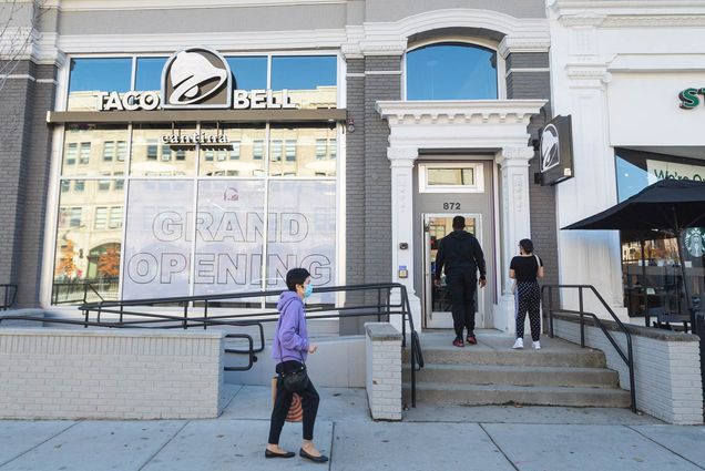 Photo of the exterior of Taco Bell on Commonwealth Ave. Two people are seen about to walk in the door, and one person in a purple sweatshirt and face mask walks by.
