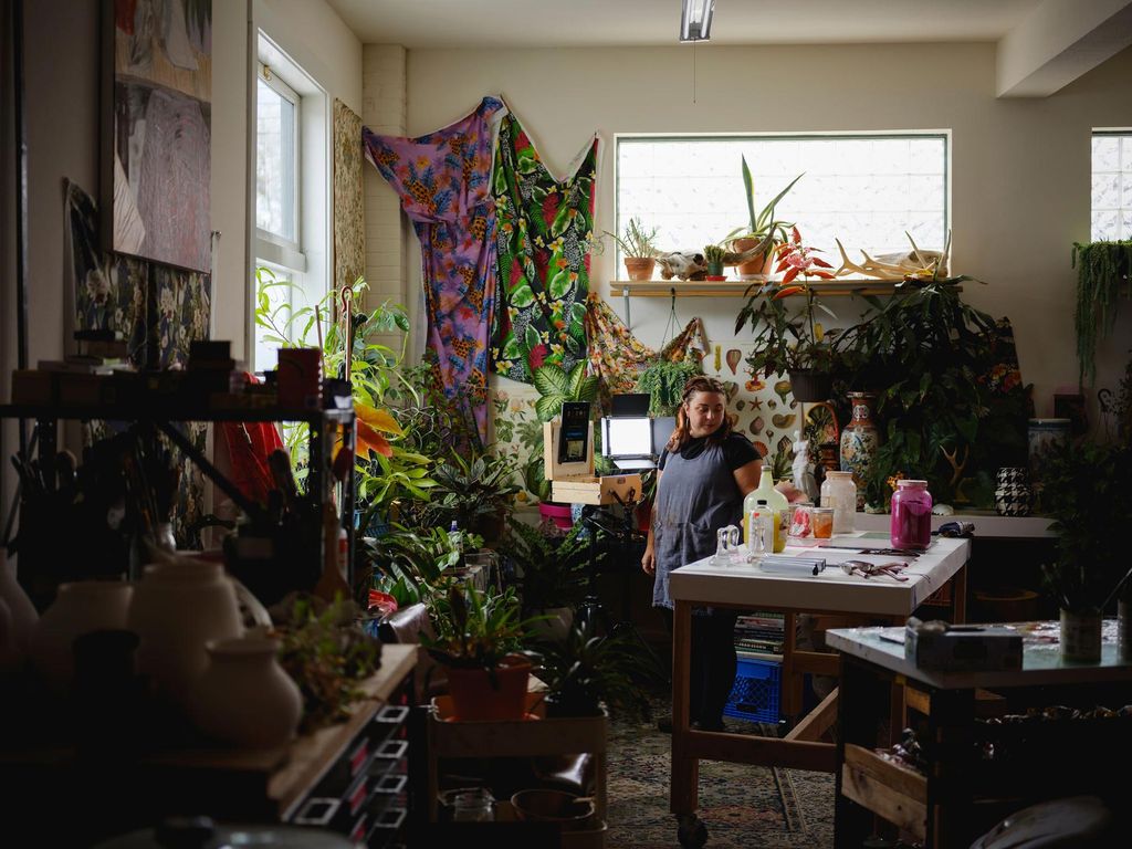 Photo of Anna Valdez (’13) in her California studio that is filed with plants. Valdez wears a gray smock and looks at jars on her table as light pours in through the window on the left. Paints and cabinets are seen in the foreground on the left.