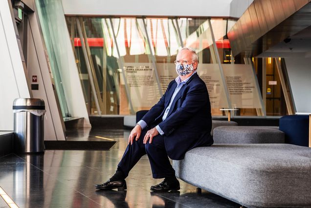 Photo of BU president Bob Brown sitting on a couch in the lobby of the Booth Theater. He wears a suit and a colorful face mask and looks toward the camera.