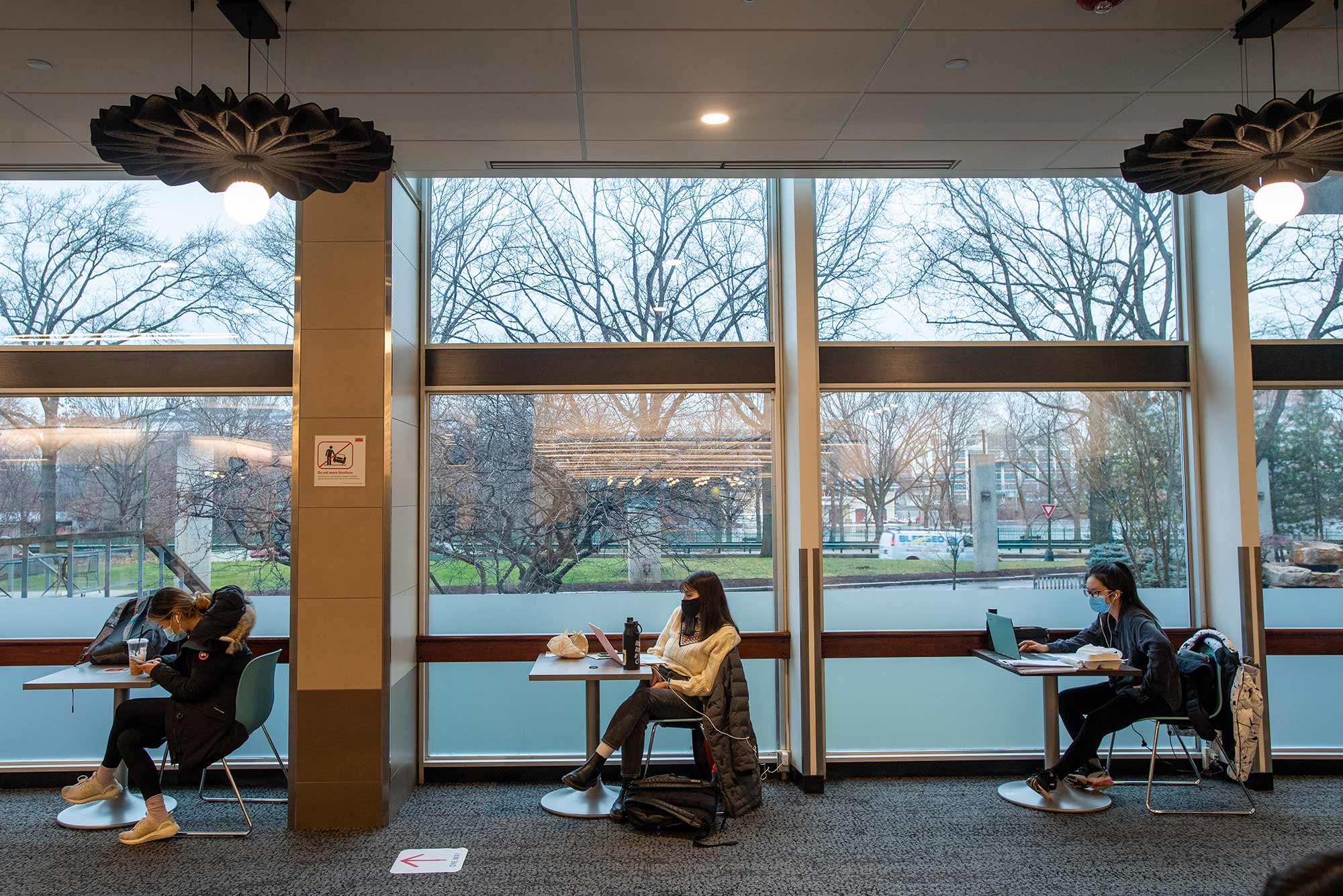 Photo of three students sitting at single person tables as they study in front of their laptops with face masks on. The bare trees of the BU Beach are seen through the windows behind them.