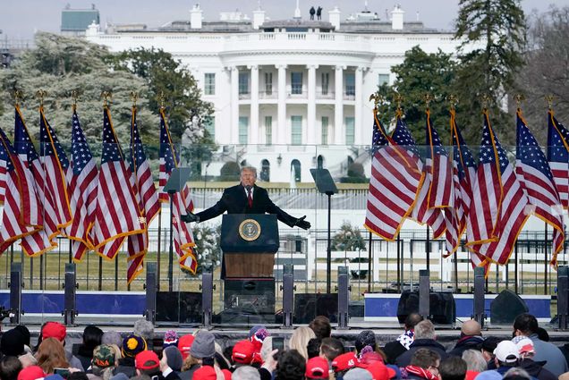 Photo of President Trump addressing supporters Wednesday January 6, 2020, falsely alleging Democrats stole November’s presidential election, before they violently breached the Capitol during the Electoral College vote recording. Trump stands behind a plexiglass barrier; the heads of people in the crowd are seen before him as he extends his arms. He is flanked onstage by American flags.