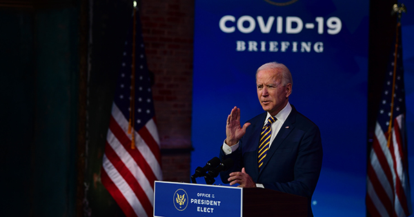 Photo from December 29, 2020 of U.S. President-elect Joe Biden delivering remarks on the ongoing coronavirus (COVID-19) pandemic at the Queen Theater on December 29, 2020 in Wilmington, Delaware. Biden stands behind a podium that reads "Office of the President Elect," his hand raised. Behind him, the words "COVID-19 Briefing" are in a white on a blue background.