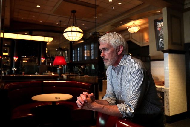 Garrett Harker, pictured in Boston on June 25, 2020, wears a blue and white striped button-down shirt and leans on the back of a red booth in one of his restaurants. He looks pensively to the left and is seen in profile.